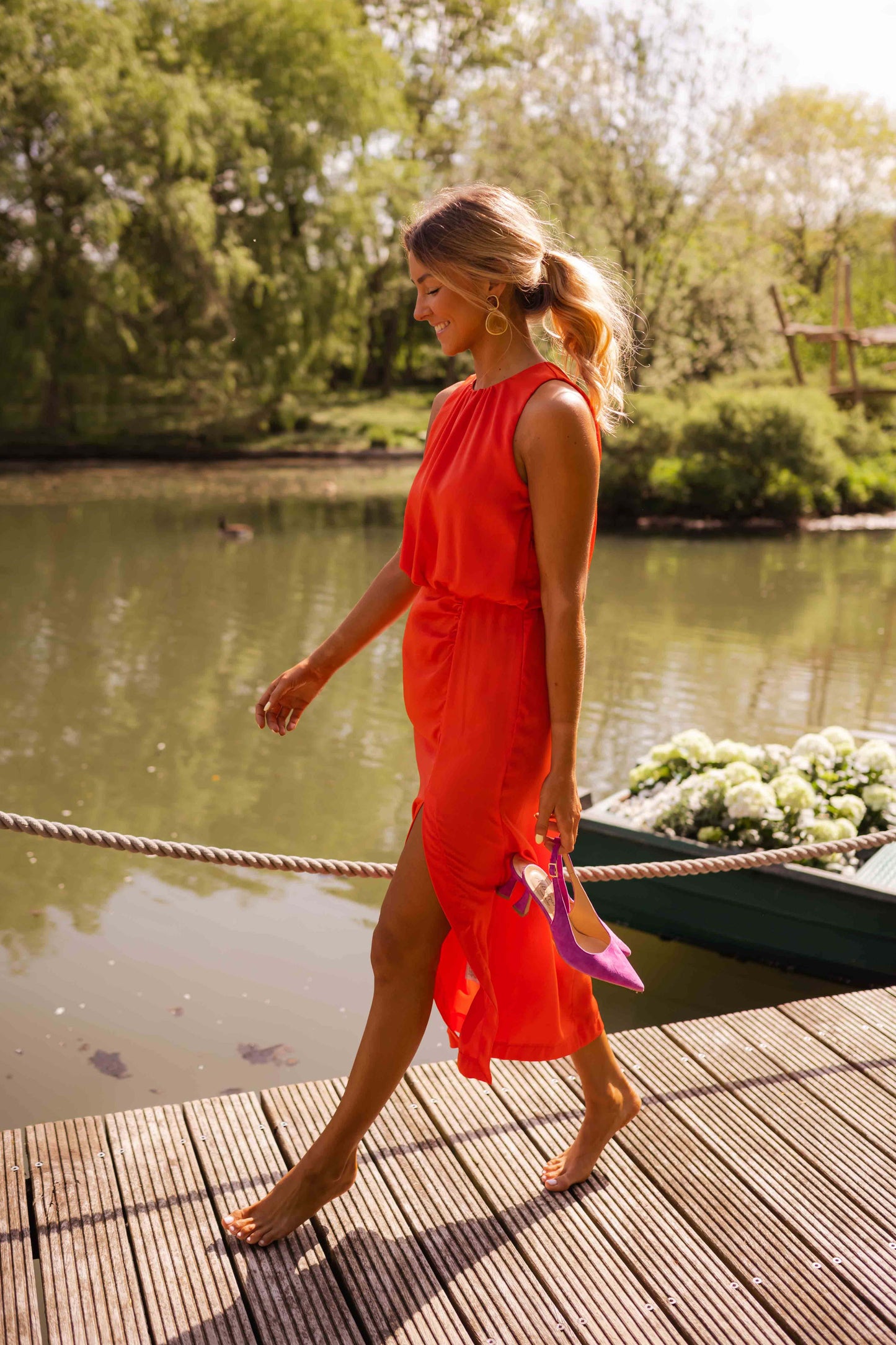 Woman in a red dress walking on a wooden dock by a body of water with greenery.