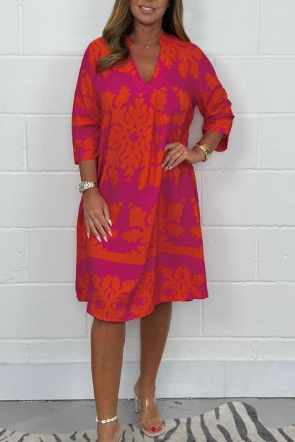 Woman wearing a vibrant pink and orange patterned dress against a white brick wall.