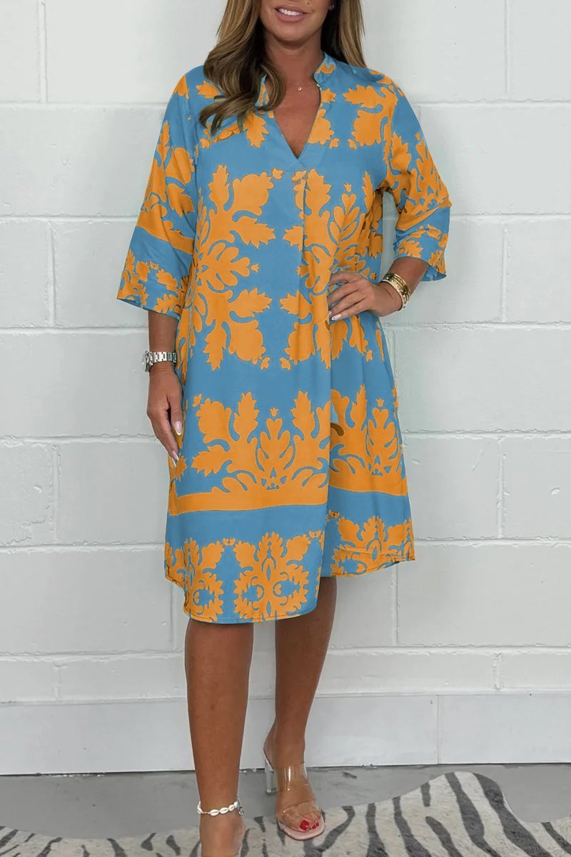 Woman wearing a blue and orange floral dress against a white brick wall.