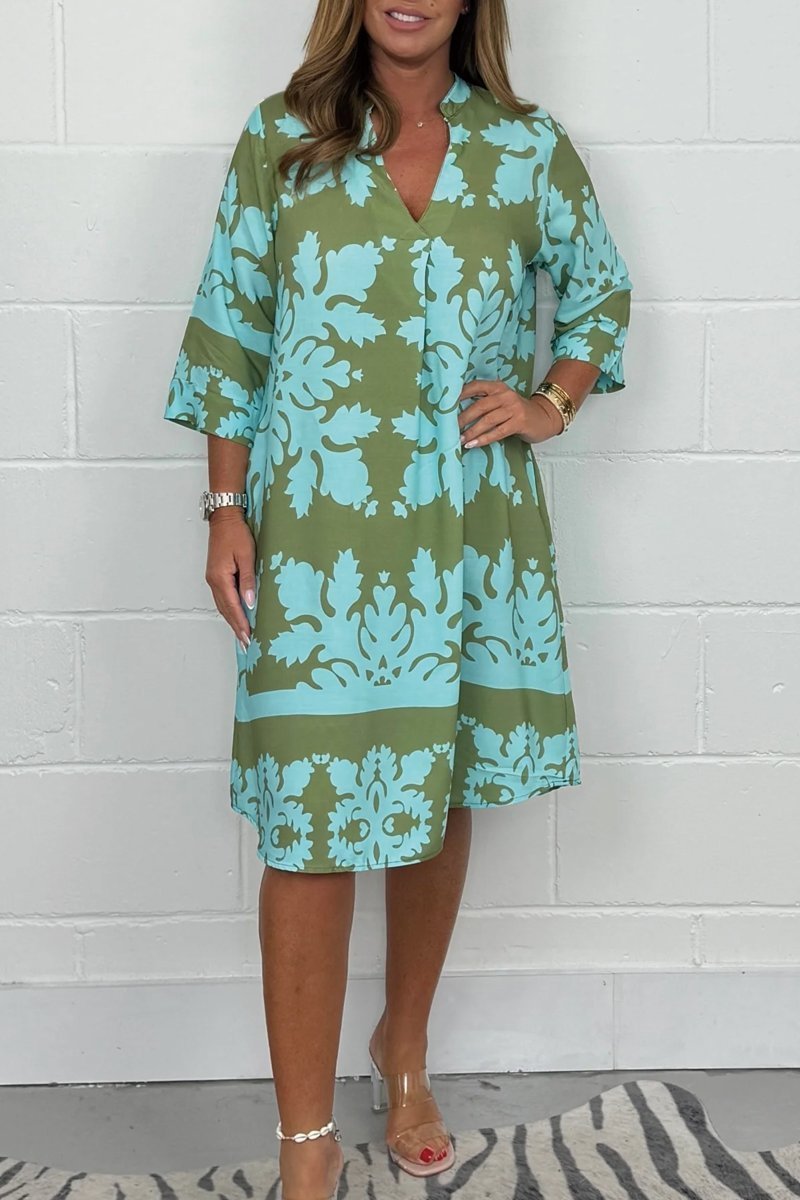 Woman wearing a green and blue patterned dress against a white brick wall.