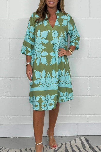 Woman wearing a green and blue patterned dress against a white brick wall.