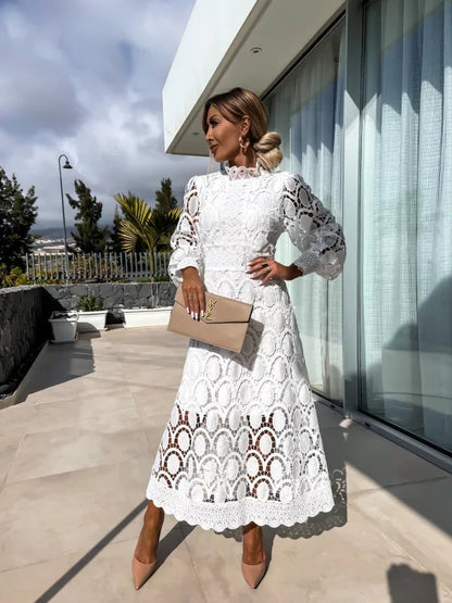 Woman in a white lace dress standing on a rooftop with modern architecture and greenery in the background.