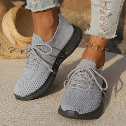 Gray knit sneakers with black soles worn by a person on a textured surface.