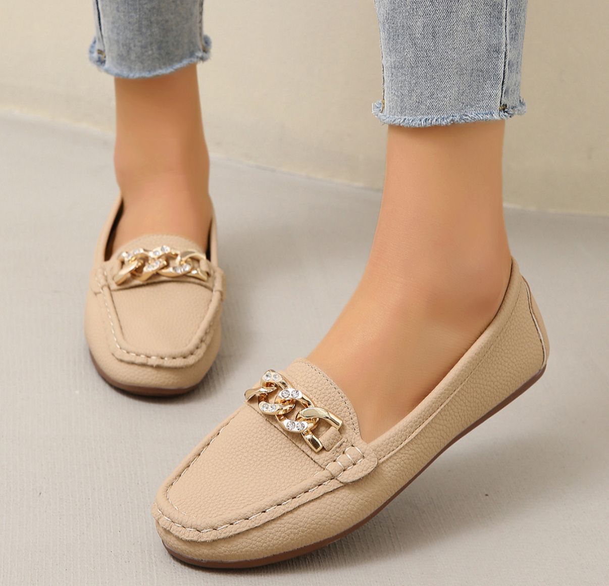 Beige loafers with gold chain details worn by a person on a neutral background