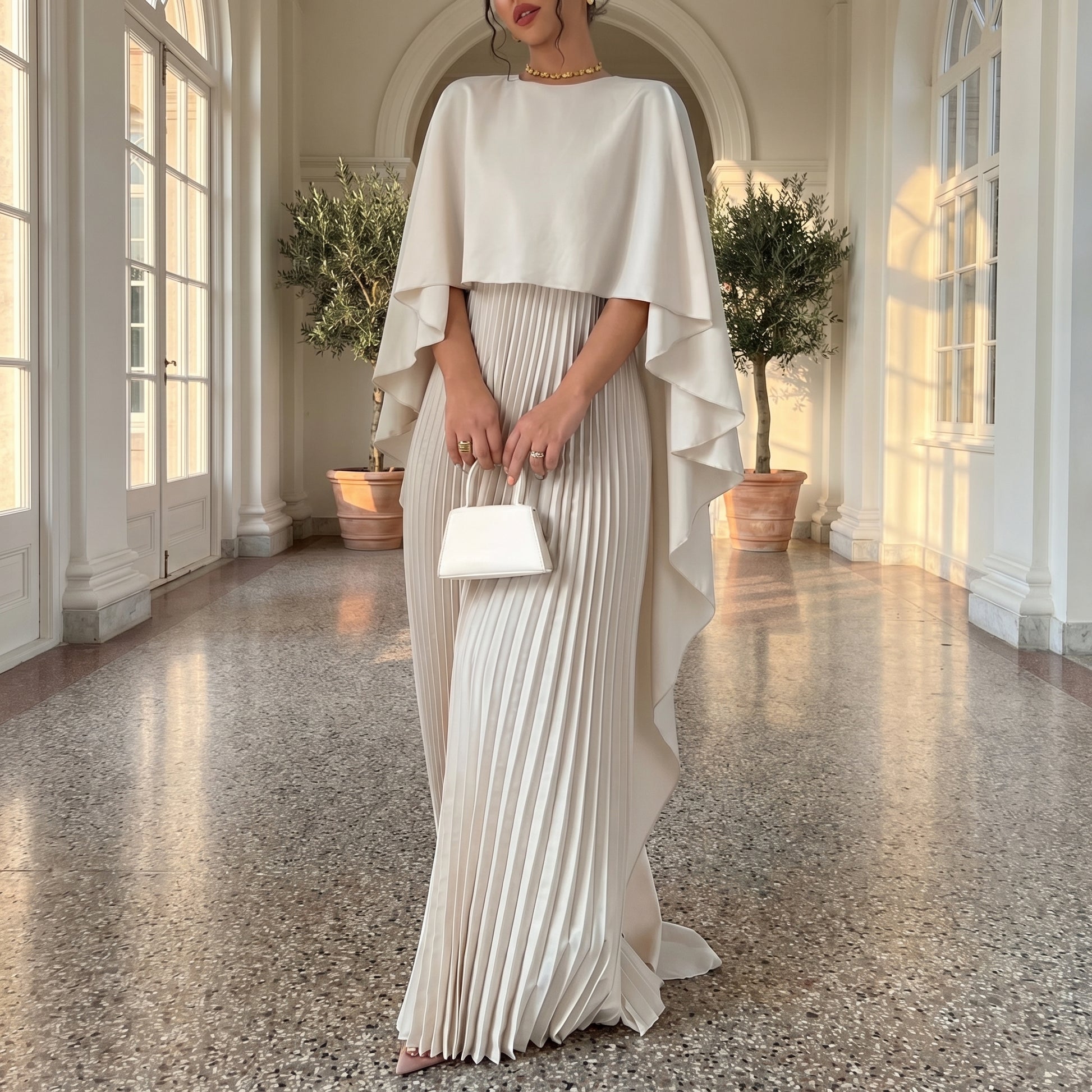 Woman in a white pleated dress with a cape in a sunlit room.