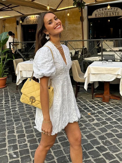 Woman in a white dress with a yellow bag walking on a street with tables and chairs in the background.