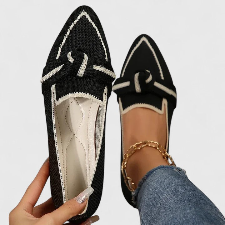 Black pointed-toe flats with white accents worn by a person on a light gray background