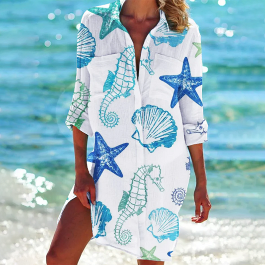 Person wearing a white cover-up with blue starfish, seashells, and seahorse patterns on a beach.