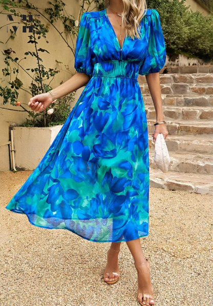 Woman wearing a blue floral dress standing outdoors on a gravel path with steps and greenery in the background.