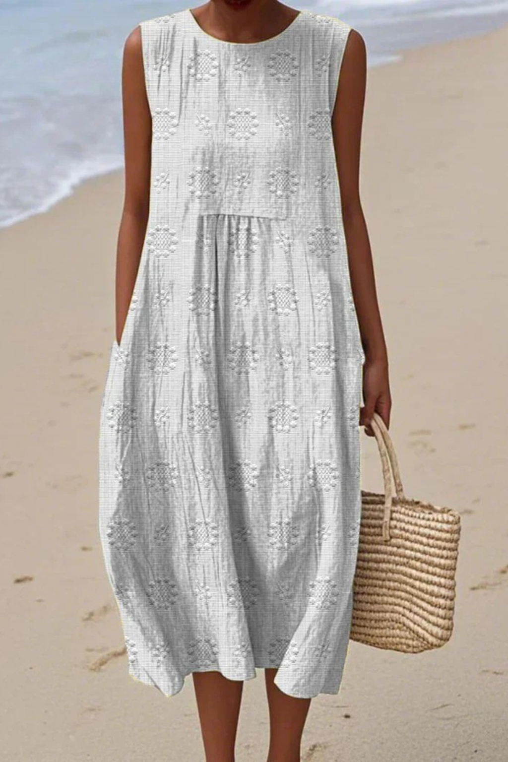 Woman in a white sleeveless dress standing on a beach with a straw bag.