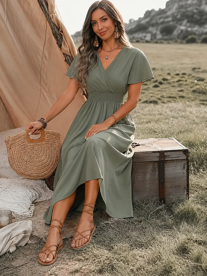 Woman in a green dress sitting on a wooden chest in a field with a tent and straw bag in the background.
