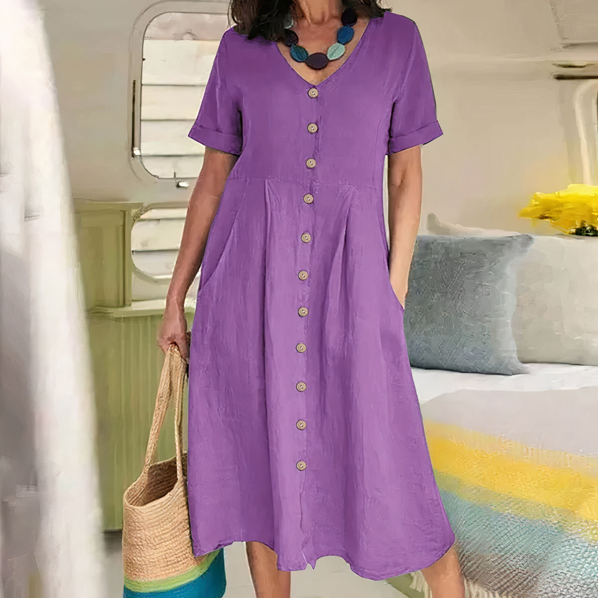 Woman wearing a purple dress holding a straw bag in a room with a bed and pillows.