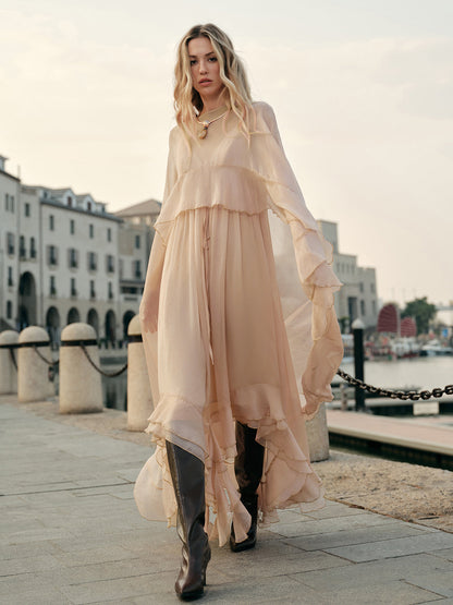 Woman in a flowing beige dress standing on a waterfront promenade with buildings in the background.