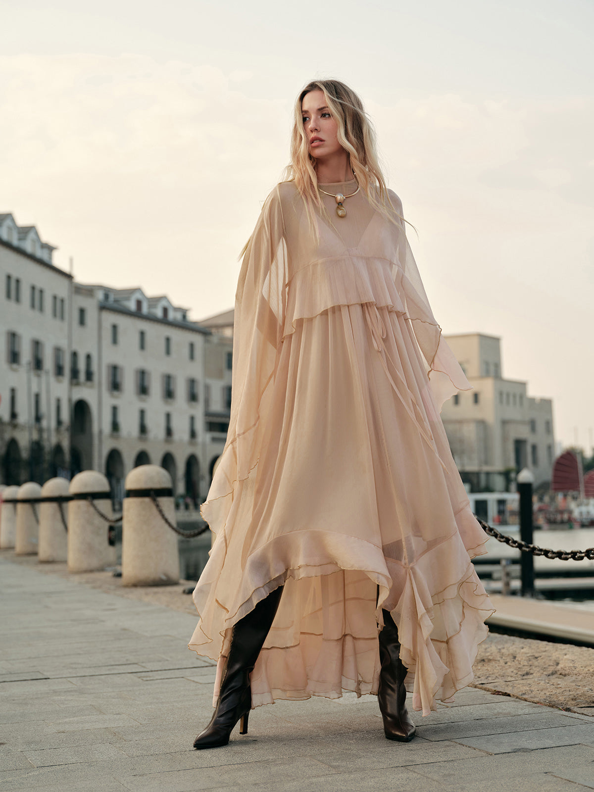 Woman in a long, light-colored dress standing on a waterfront with buildings in the background.