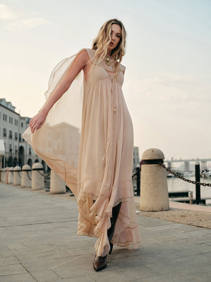 Woman in a flowing beige dress standing on a waterfront promenade.