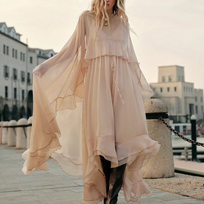 Woman in a flowing beige dress standing on a waterfront promenade with buildings in the background.