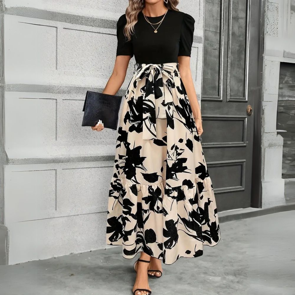 Woman wearing a black top and floral skirt with a clutch in front of a gray door.
