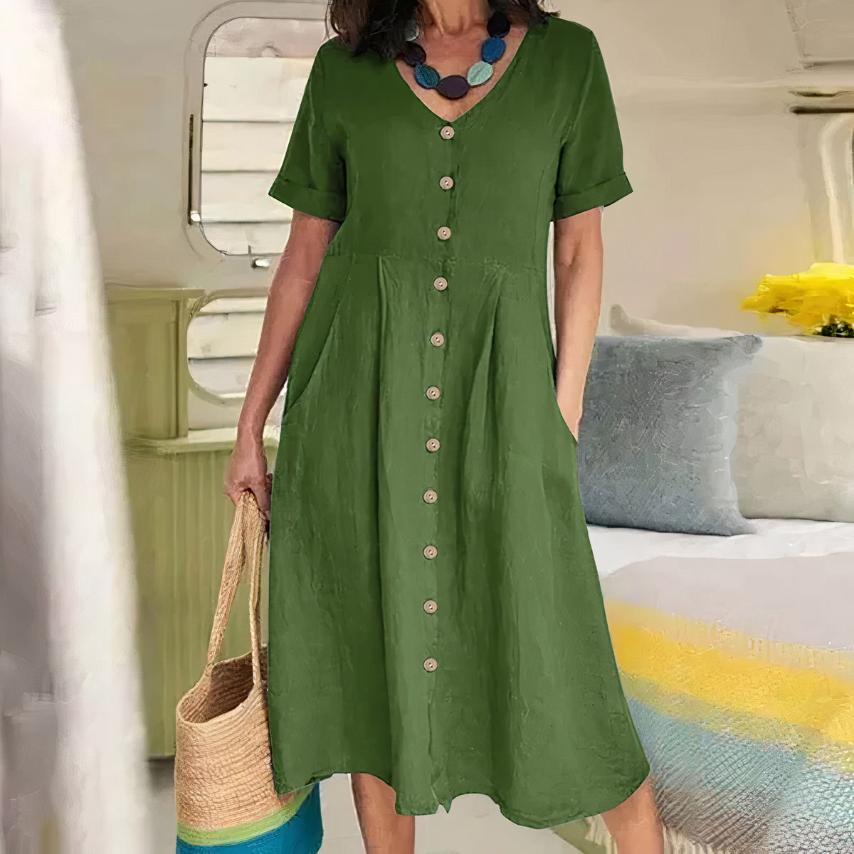 Woman wearing a green dress with buttons, holding a straw bag, in a room with a bed and pillows.