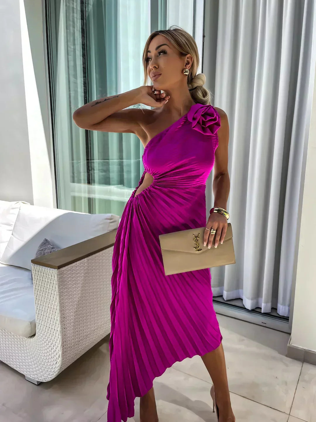 Woman in a vibrant pink dress holding a beige clutch in a room with a bed and large window.