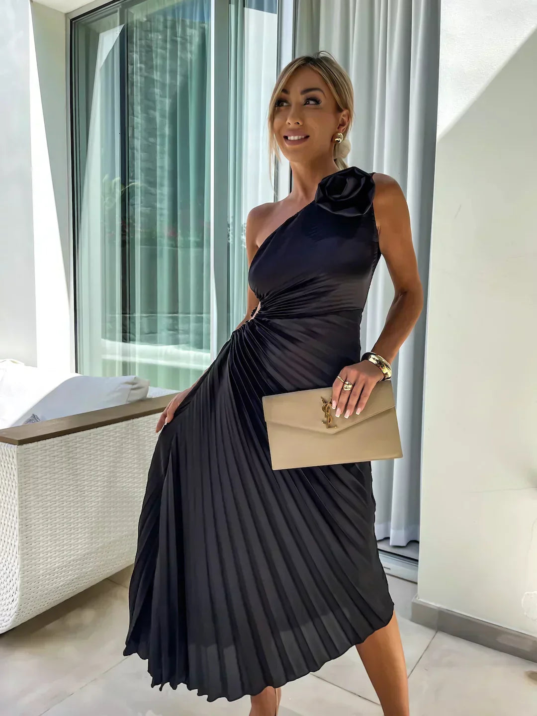 Woman in a black one-shoulder dress with a floral detail, holding a beige clutch, standing in a modern indoor setting.
