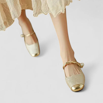 Gold ballet flats with pearl accents worn with a light-colored skirt on a white background