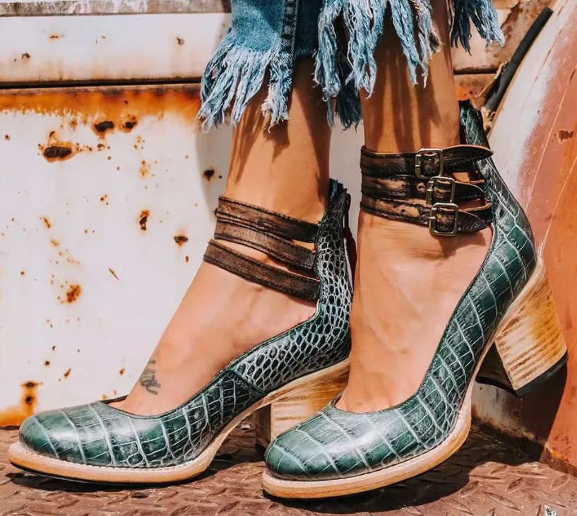 Green crocodile-patterned shoes with brown straps worn by a person against a rusty metal background.