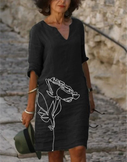Woman wearing a black dress with white floral design, standing against a stone wall.