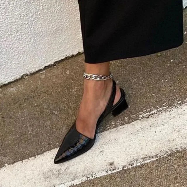 Black pointed-toe shoes worn with a chain ankle strap on a sidewalk.