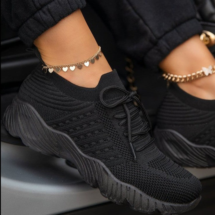 Black sneakers worn with gold anklets on a neutral background