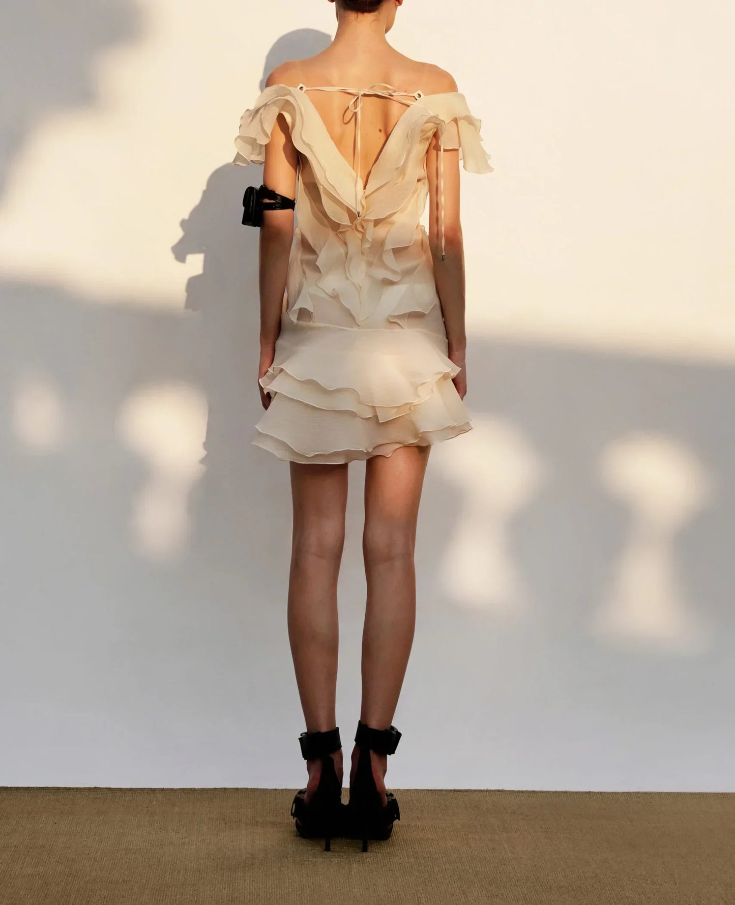 Woman wearing a beige ruffled dress with black shoes against a white wall.