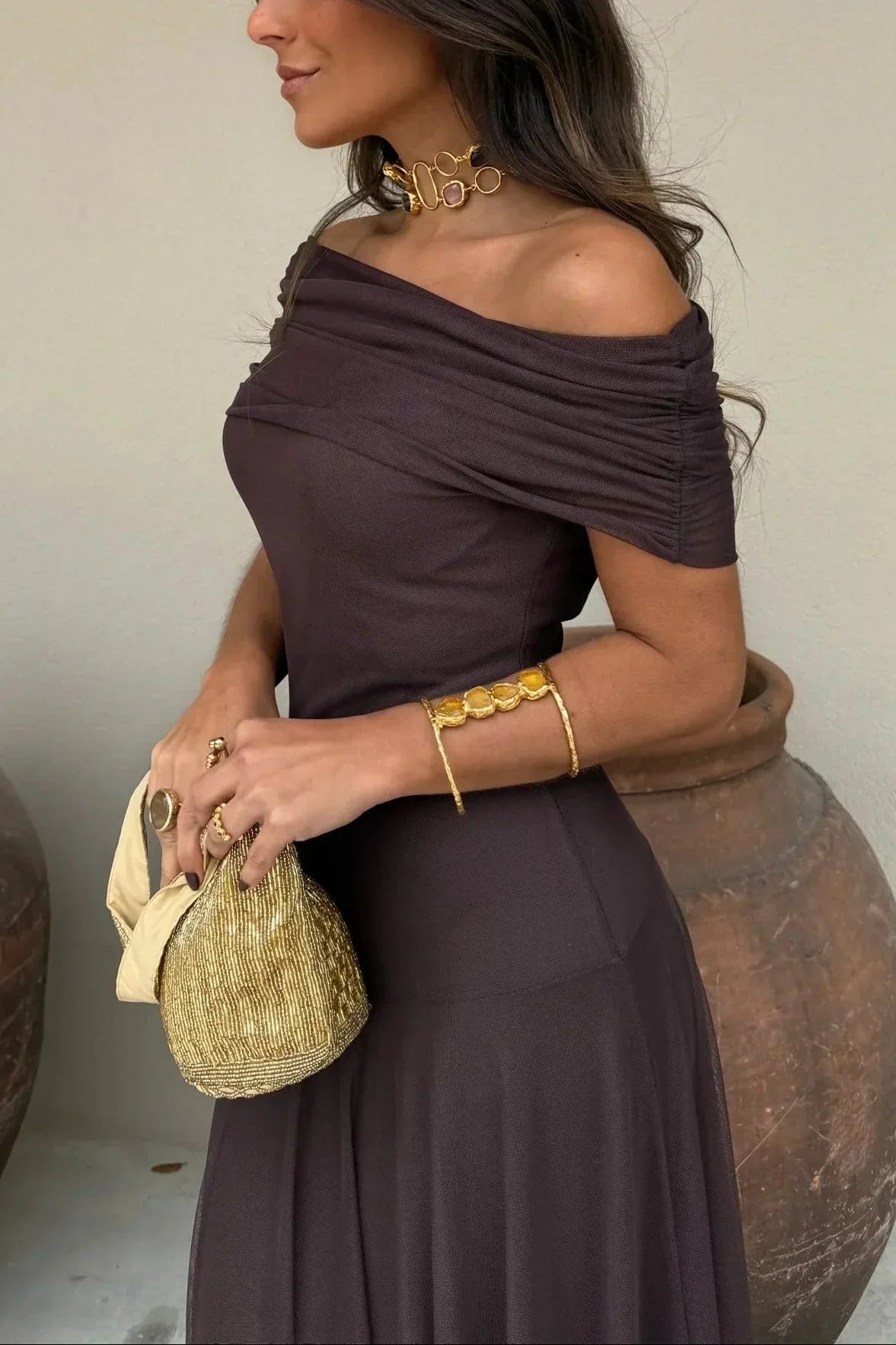 Woman in a brown off-shoulder dress holding a beige handbag, wearing gold jewelry.