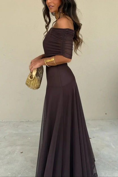 Woman in a dark brown off-shoulder dress holding a gold clutch against a plain background