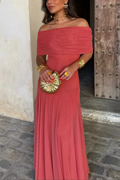 Woman in a coral off-shoulder dress holding a gold clutch against a stone wall.