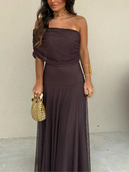 Woman wearing a strapless brown dress holding a gold handbag against a plain background