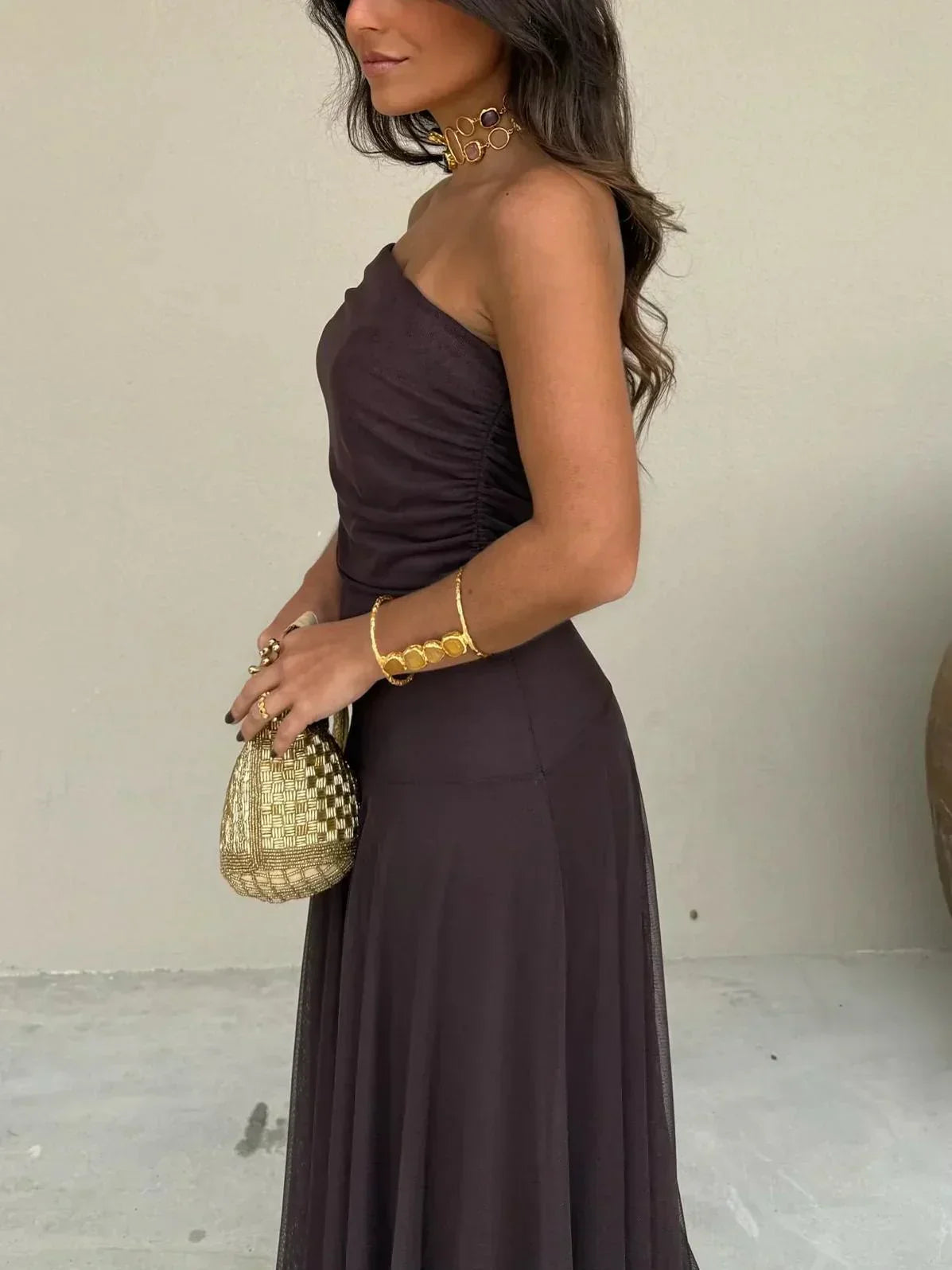 Woman in a dark brown one-shoulder dress holding a gold handbag against a neutral background