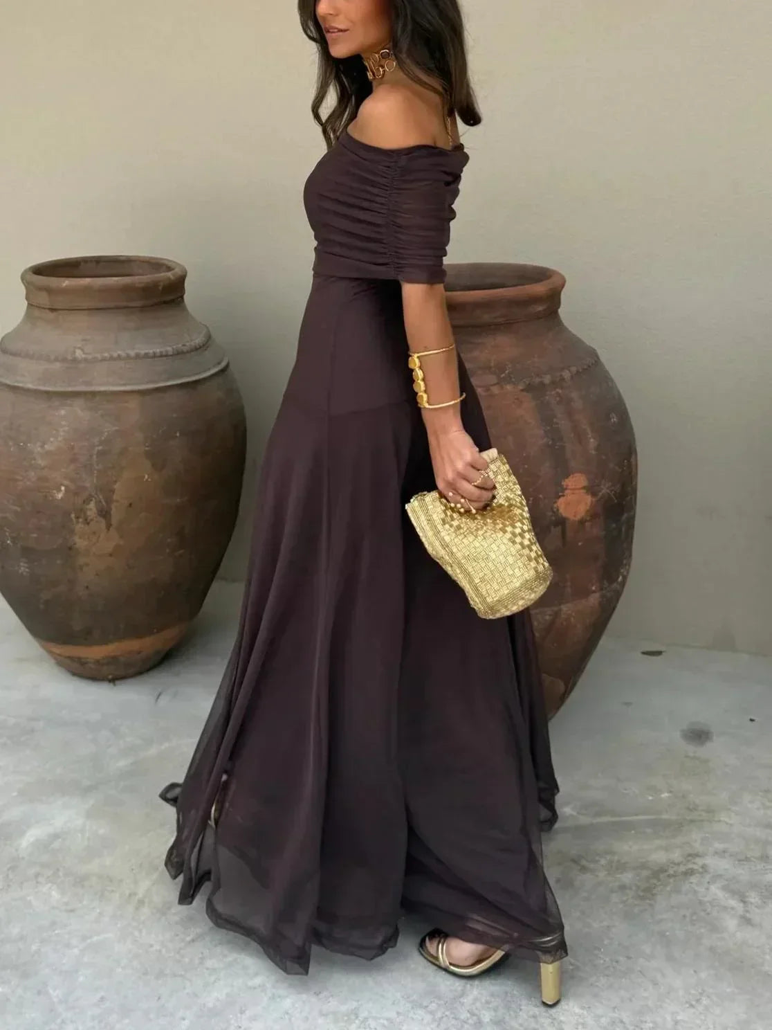 Woman in a long dark purple dress holding a gold clutch against a neutral background with large pots.