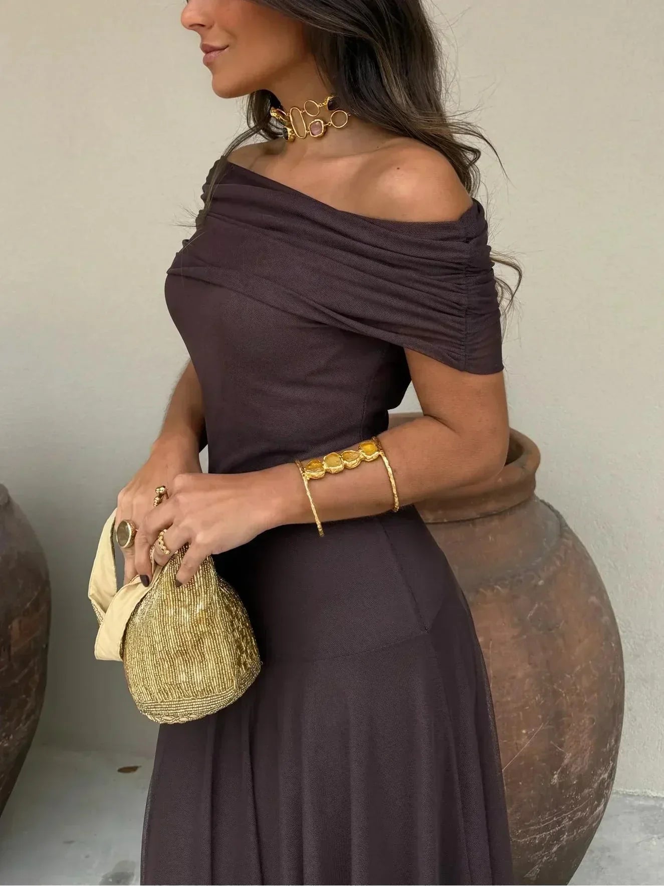 Woman in a dark brown off-shoulder dress holding a gold clutch, standing against a neutral background.