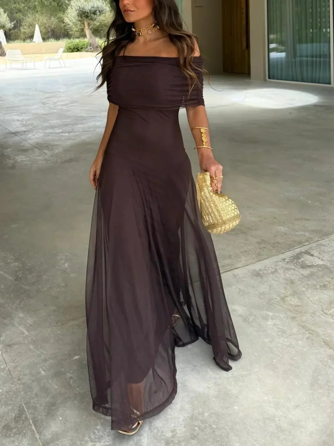 Woman in a long brown dress holding a gold purse on a stone patio.