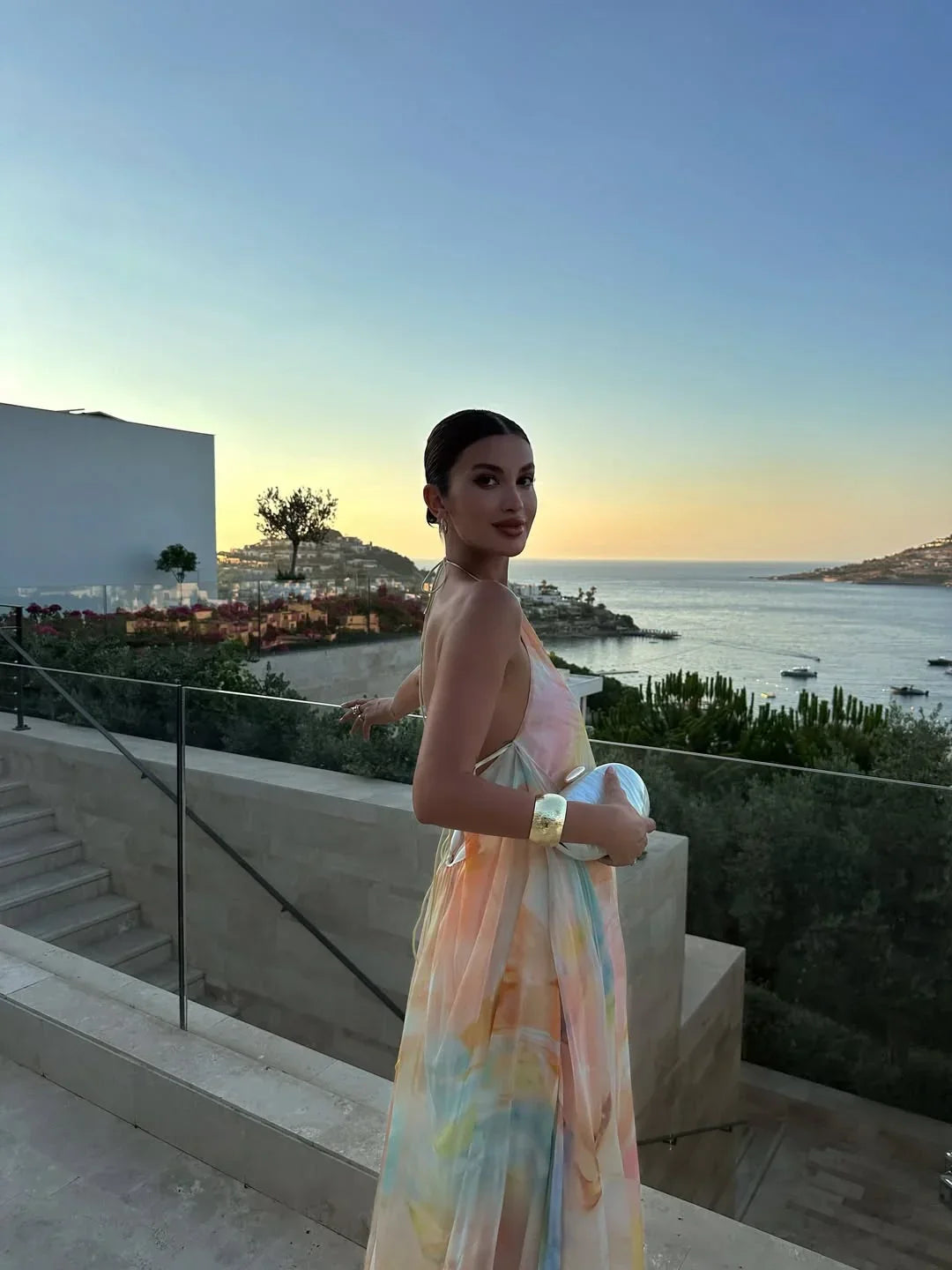 Woman in a colorful dress standing on a balcony overlooking a coastal sunset.