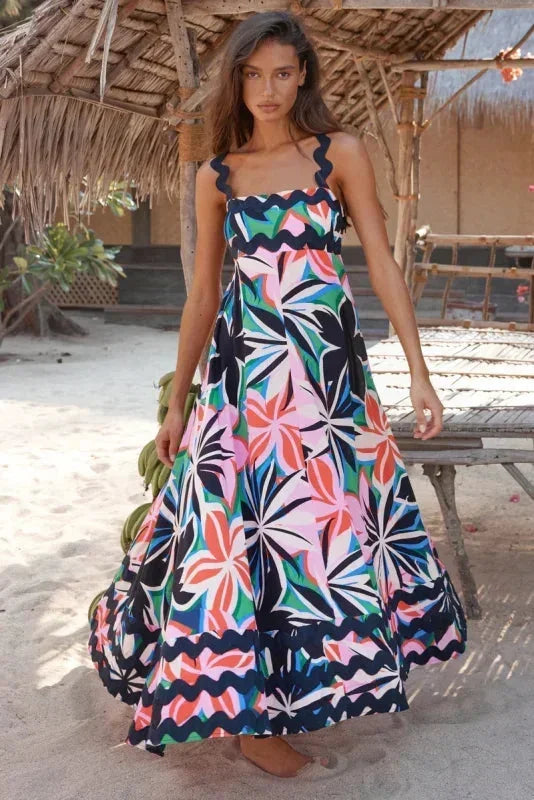 Woman wearing a colorful floral dress standing on a sandy beach with a thatched roof structure in the background.