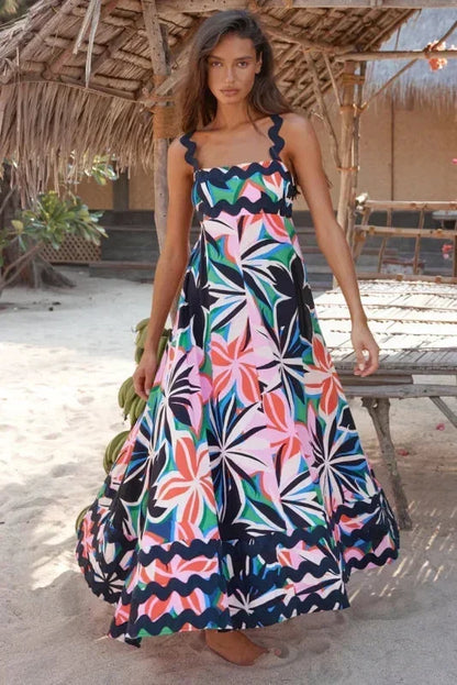 Woman wearing a colorful floral dress standing on a sandy beach with a thatched roof structure in the background.