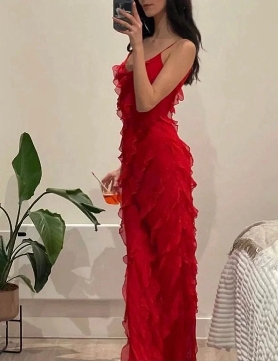 Woman in a red ruffled dress holding a drink, standing in a room with a plant and chair.