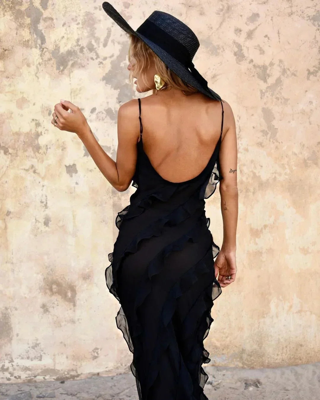 Woman wearing a black dress with a wide-brimmed hat against a textured wall.