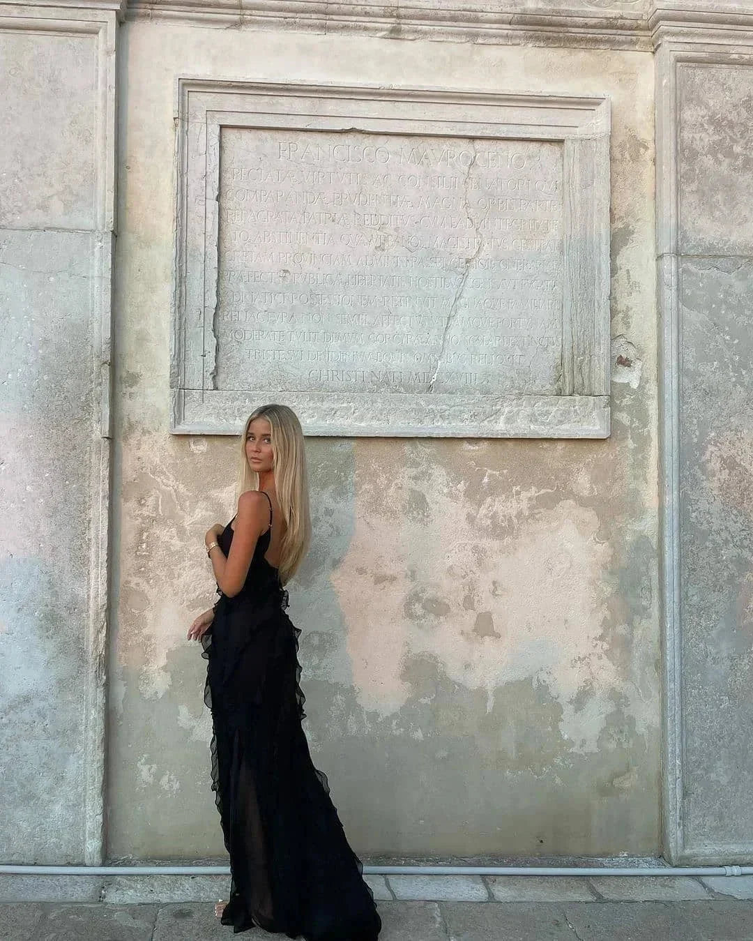 Woman in a black dress standing against a textured stone wall.