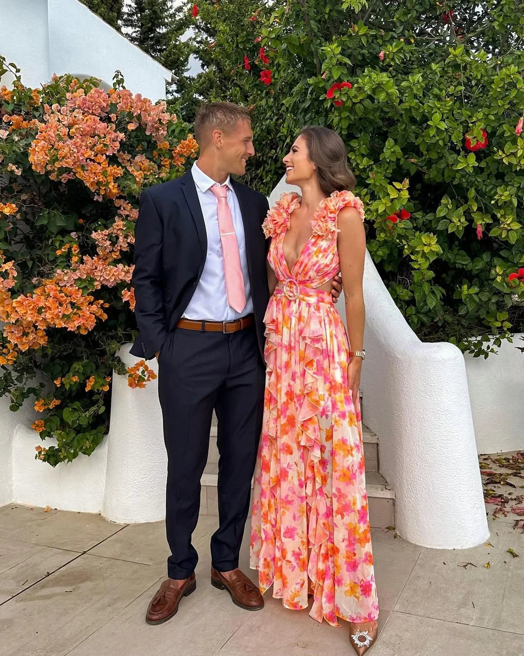 Man in a suit and woman in a floral dress standing together outdoors with flowers and greenery in the background.