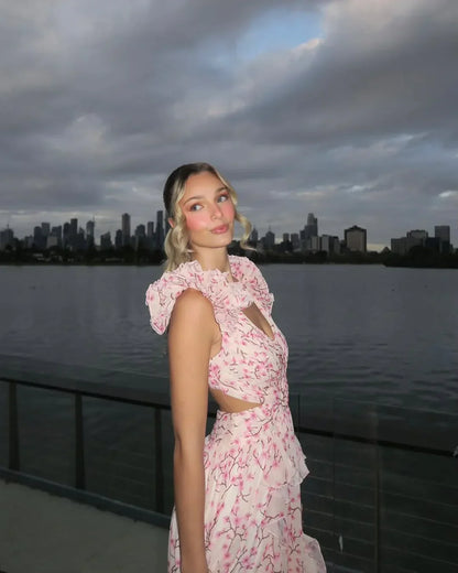 Woman in a pink floral dress standing by a waterfront with a city skyline in the background