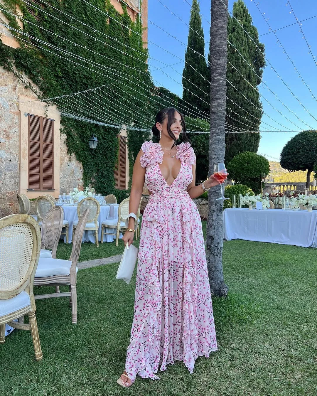 Woman in a pink floral dress standing outdoors at a formal event with tables and chairs.