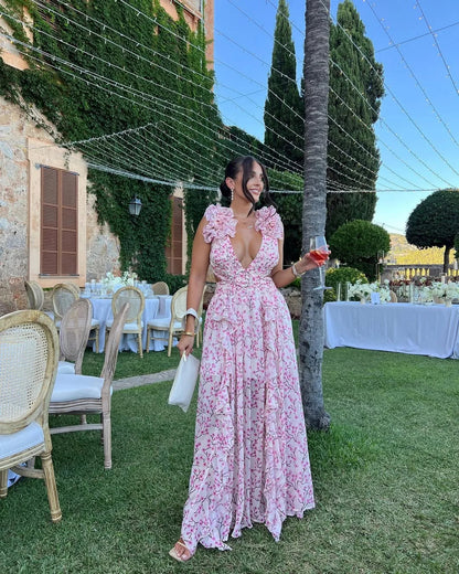 Woman in a pink floral dress standing outdoors at a formal event with tables and chairs.