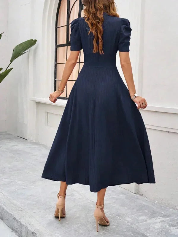 Woman wearing a navy blue dress standing on a stone path with a white wall and plant in the background.