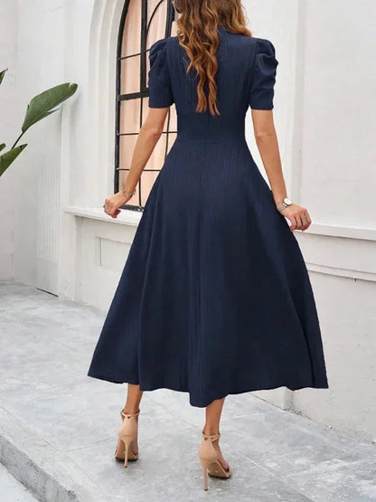 Woman wearing a navy blue dress standing on a stone path with a white wall and plant in the background.